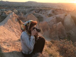 Frau sitzt auf einem Berg, guckt durch eine Kamera, scheint zu fotografieren. Im Hintergrund sind Hügelspitzen und ein Sonnenuntergang zu sehen