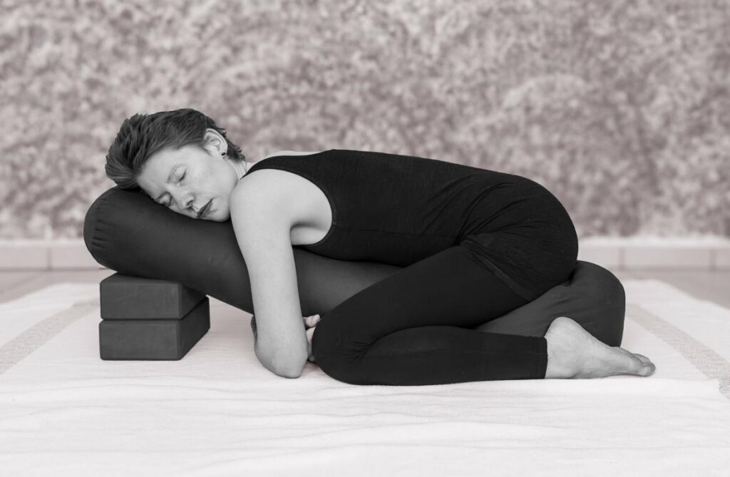 Restorative Yoga Kindspose, Frau liegt auf einem angeschrägten Bolster mit geschlossenen Augen.