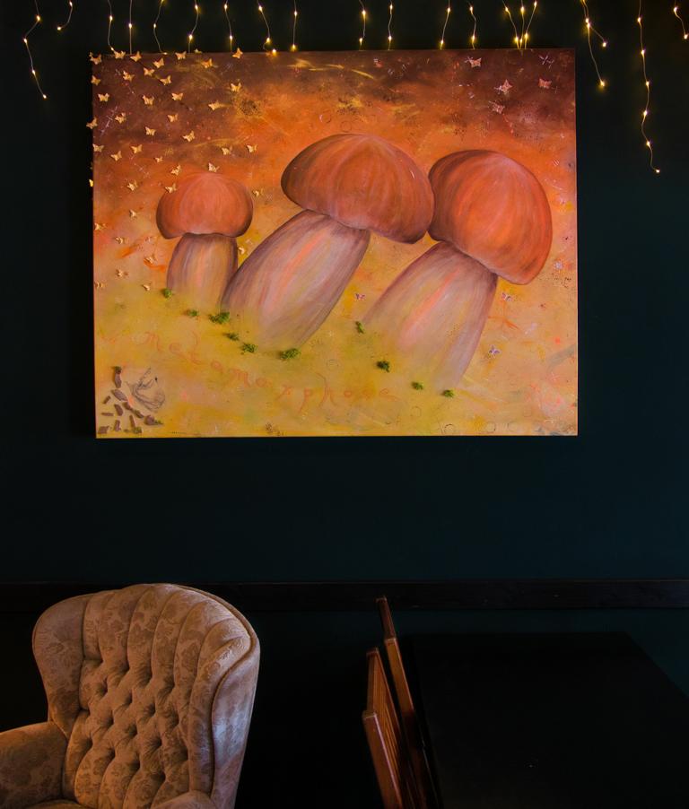 Ein Kunstwerk, in gelb-orange-braun Tönen gehalten mit Pilzen drauf, hängt an einer Wand in einer Bar